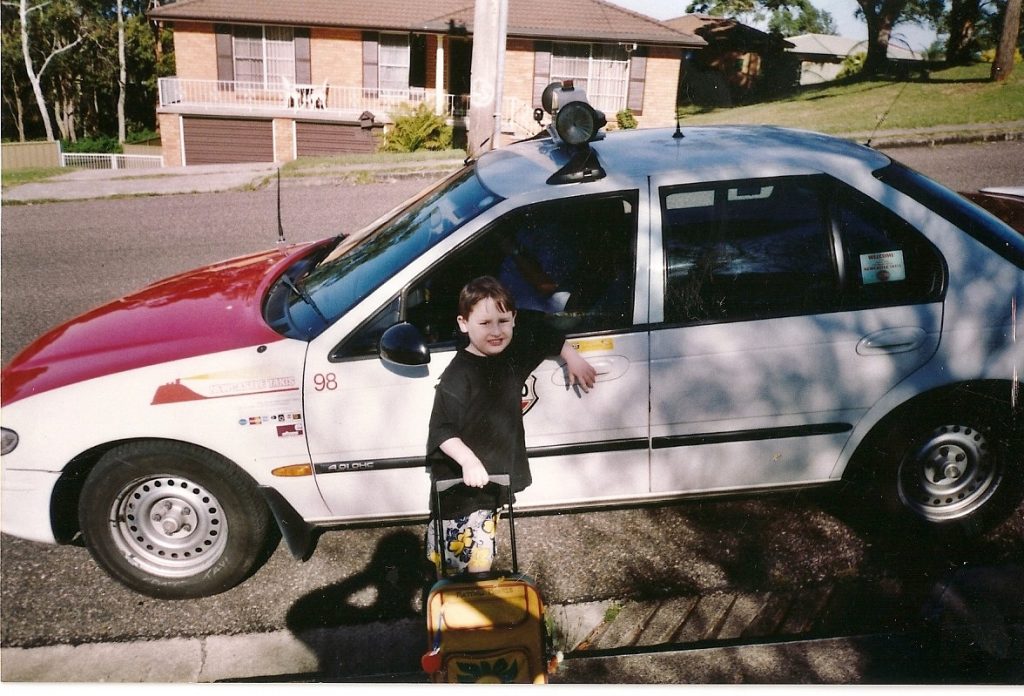 Matt stands next to a taxi, the taxi is from the 90s and has a red hood, the branding of the car reads 'Newcastle Taxis.' Matt has his arm against the passenger door of the car, a driver in a blue shirt can be seen inside, although we can't see his face.